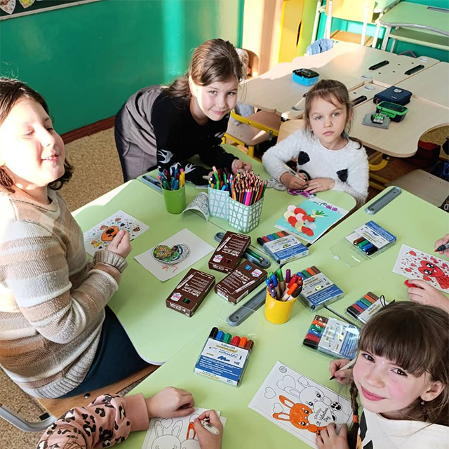 Kids excitedly holding up marker boxes and showing off their artwork in a bright classroom.